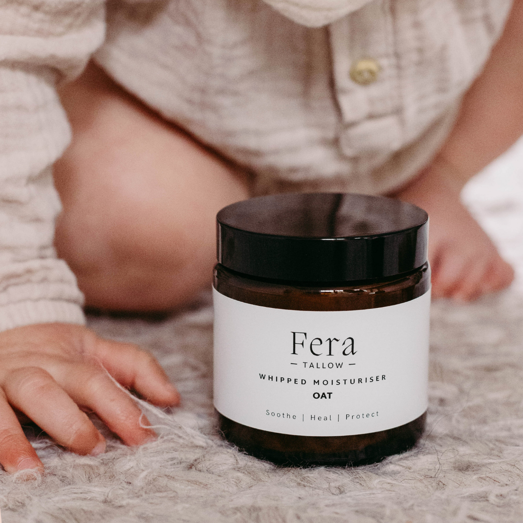 Fera Tallow whipped moisturiser jar on a textured surface with a child's hand and legs in the background.